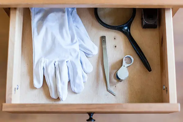 Ein Paar weißer Handschuhe aus Baumwolle liegt zusammen mit einer Pinzette und einer Lupe in einer offenen Schublade.