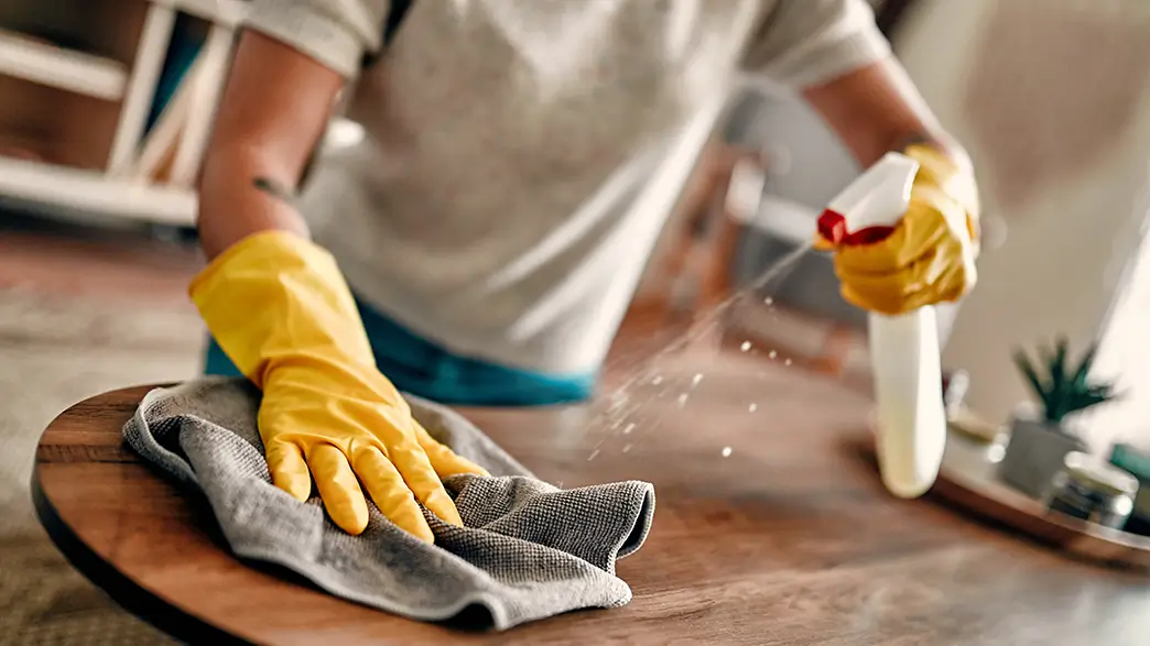 A woman wipes the surface of a wooden table with a damp cloth, using a spray bottle with cleaning solution and wearing protective gloves