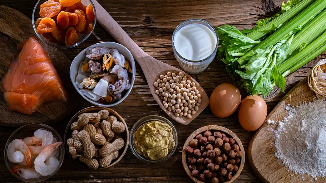 Various known food allergens are displayed on a table: fish fillet, shrimp, shellfish, peanuts, mustard, milk, eggs, seeds, wheat flour, wheat noodles, and leeks