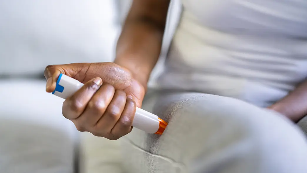 A woman in white clothing demonstrates the use of an epinephrine injector, which injects adrenaline directly into the thigh muscle during an allergic shock